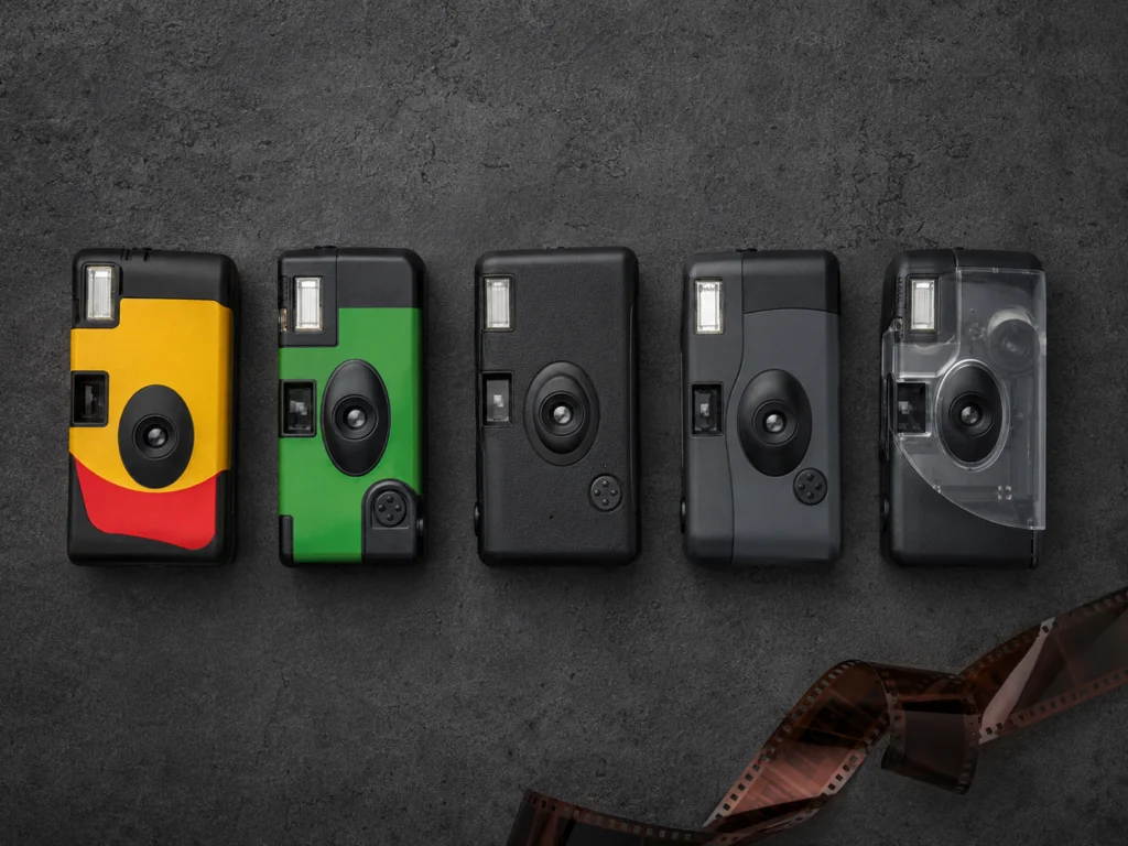 Five disposable cameras lined up on a dark surface, including Kodak FunSaver, Fujifilm QuickSnap, and Ilford single-use cameras