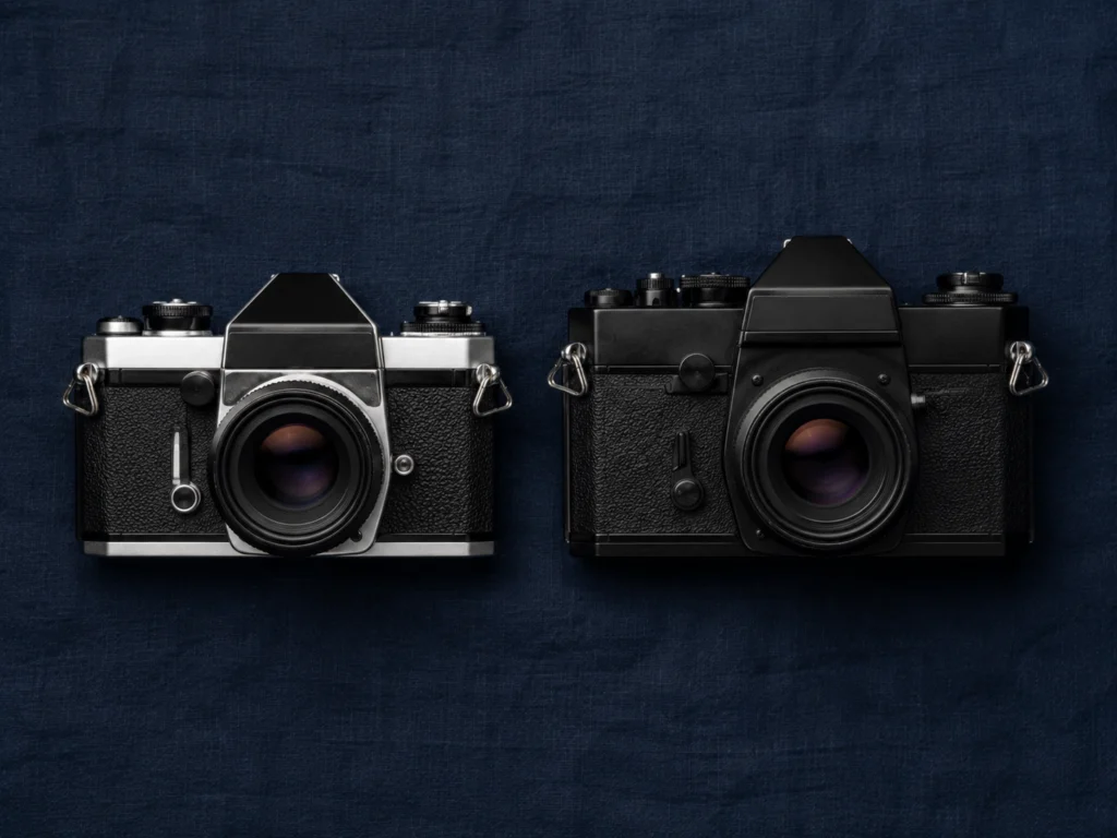 Two vintage 35mm SLR cameras placed side by side on a dark surface showing different body styles and sizes
