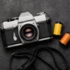 35mm SLR film camera placed next to two rolls of 35mm color film on a dark textured surface