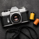 35mm SLR film camera placed next to two rolls of 35mm color film on a dark textured surface