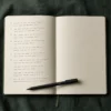 Open journal on a dark desk with a handwritten list of questions visible on the page, pen resting beside it